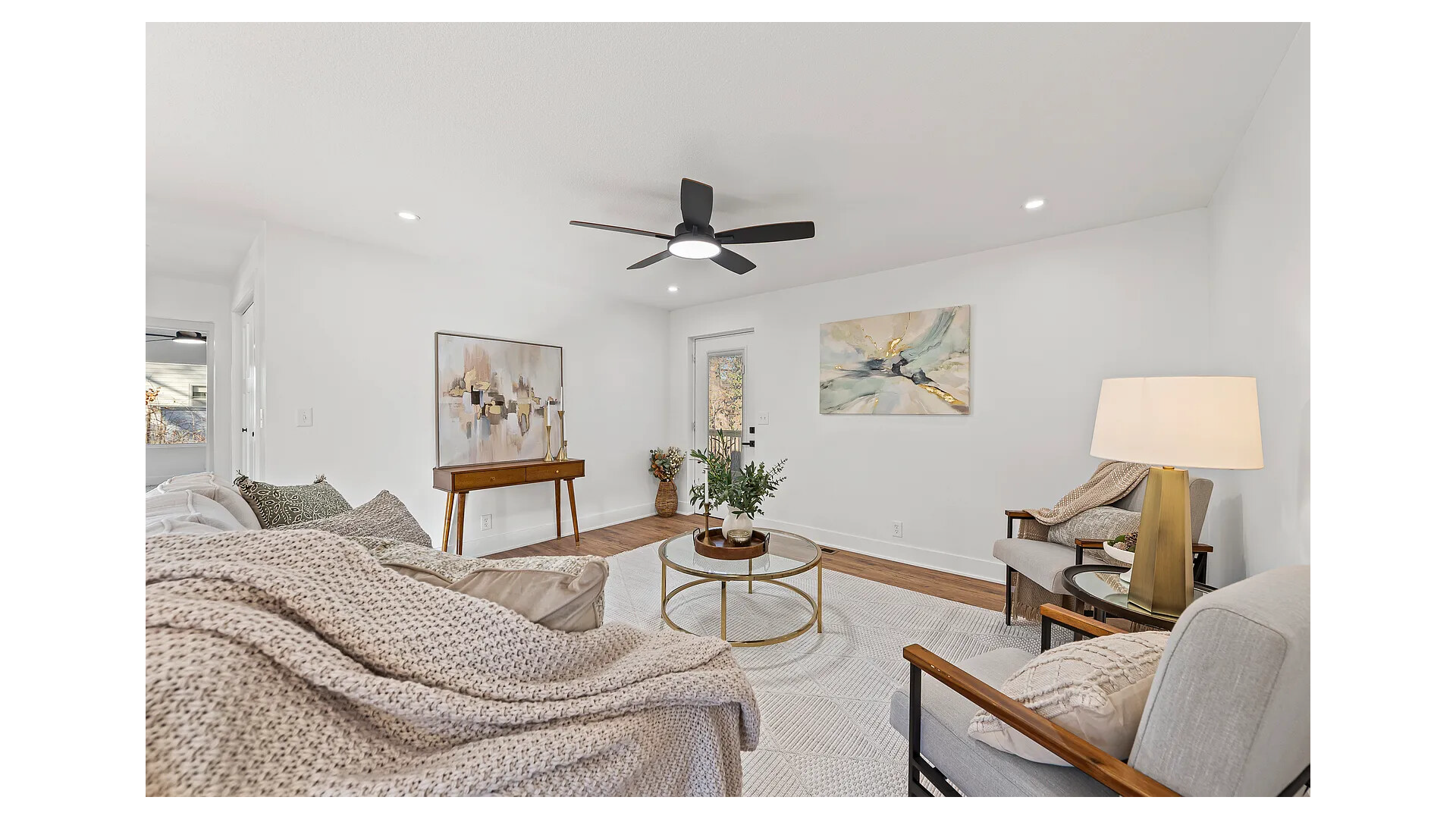 Bright living room with white walls, wooden flooring, modern furniture, a round glass coffee table with plants, on the wall are two abstract paintings, ceiling fan with black blades, and a large window letting in natural light.