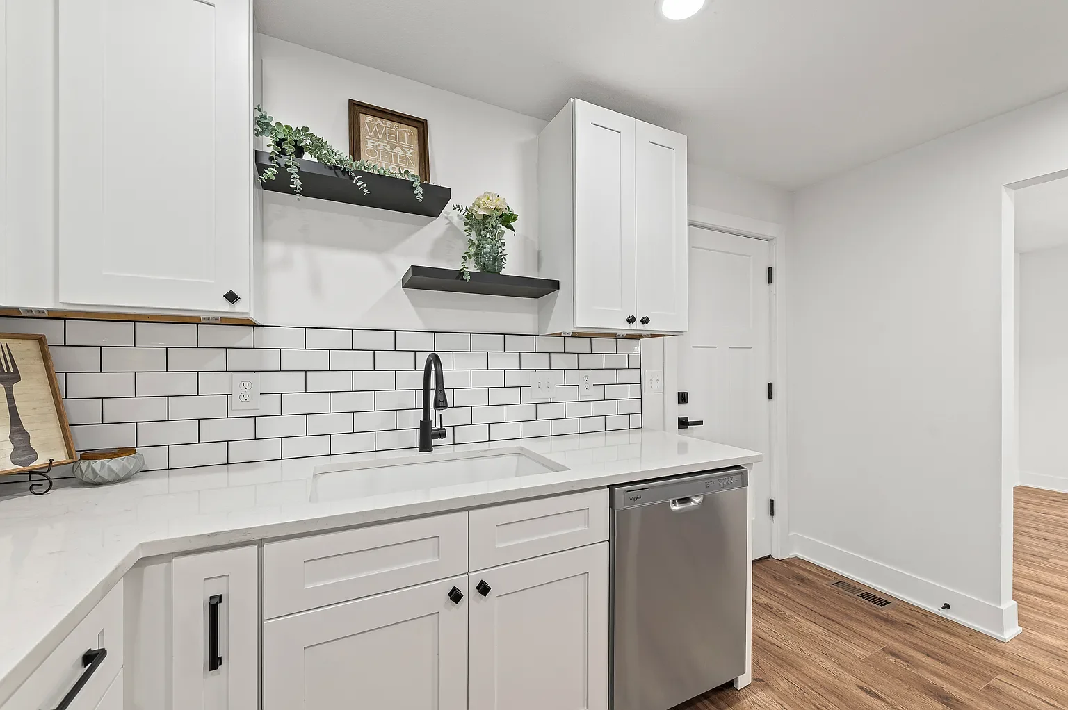Modern kitchen with white cabinets, white countertops, black hardware, black faucet, white subway tile backsplash, and stainless steel dishwasher. Decor includes black shelves, a framed quote, and plants.
