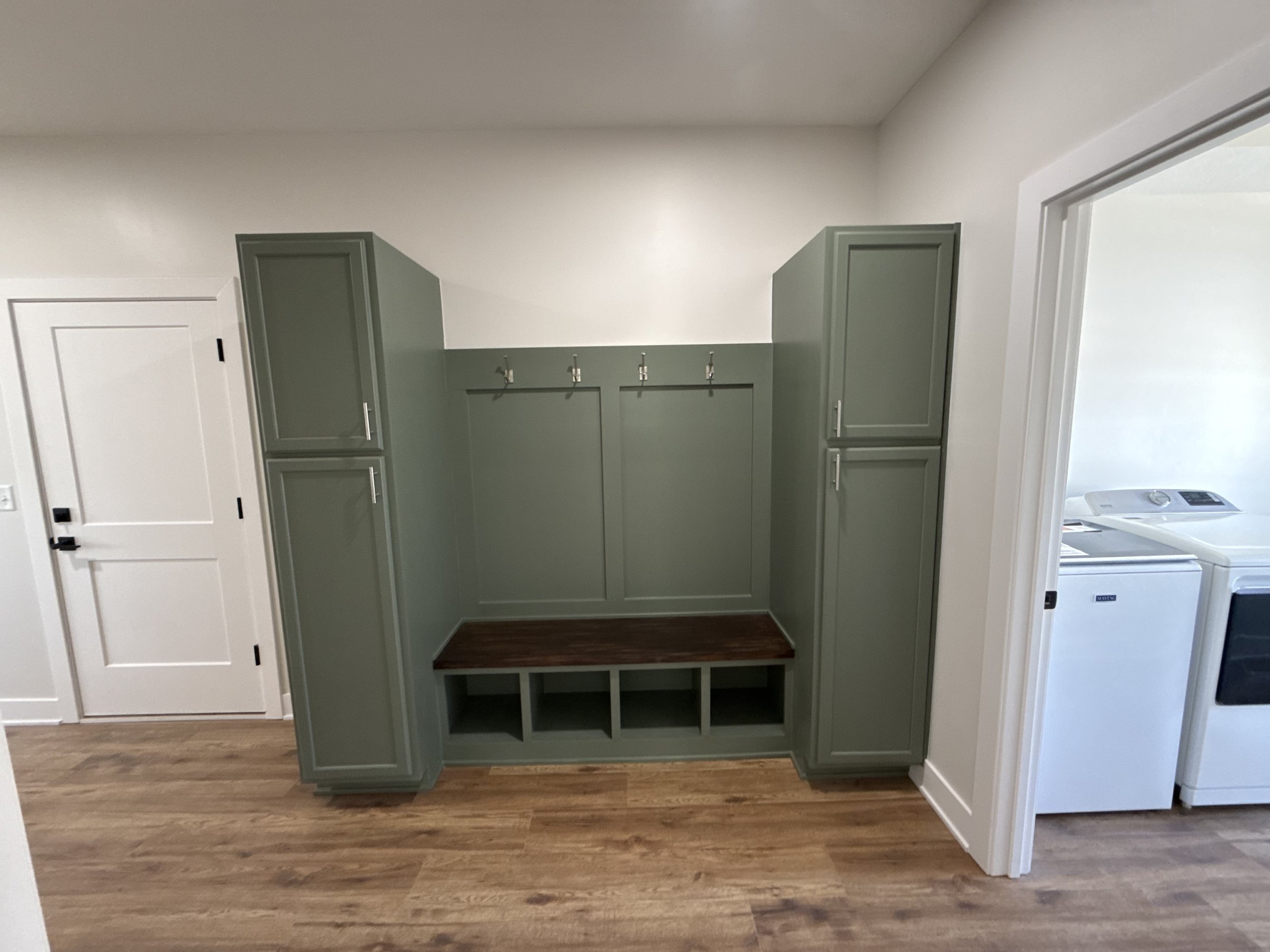 Entryway mudroom area with built-in green cabinetry, hooks for hanging items, a wooden bench with cubbies underneath, and white walls with hardwood flooring.