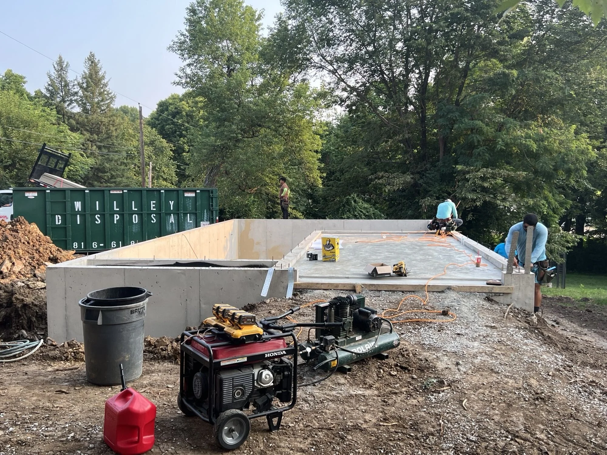 Construction site with workers installing a concrete foundation, construction tools and equipment, and a large green dumpster labeled 'WILLEY DISPOSAL' in the background, surrounded by trees.