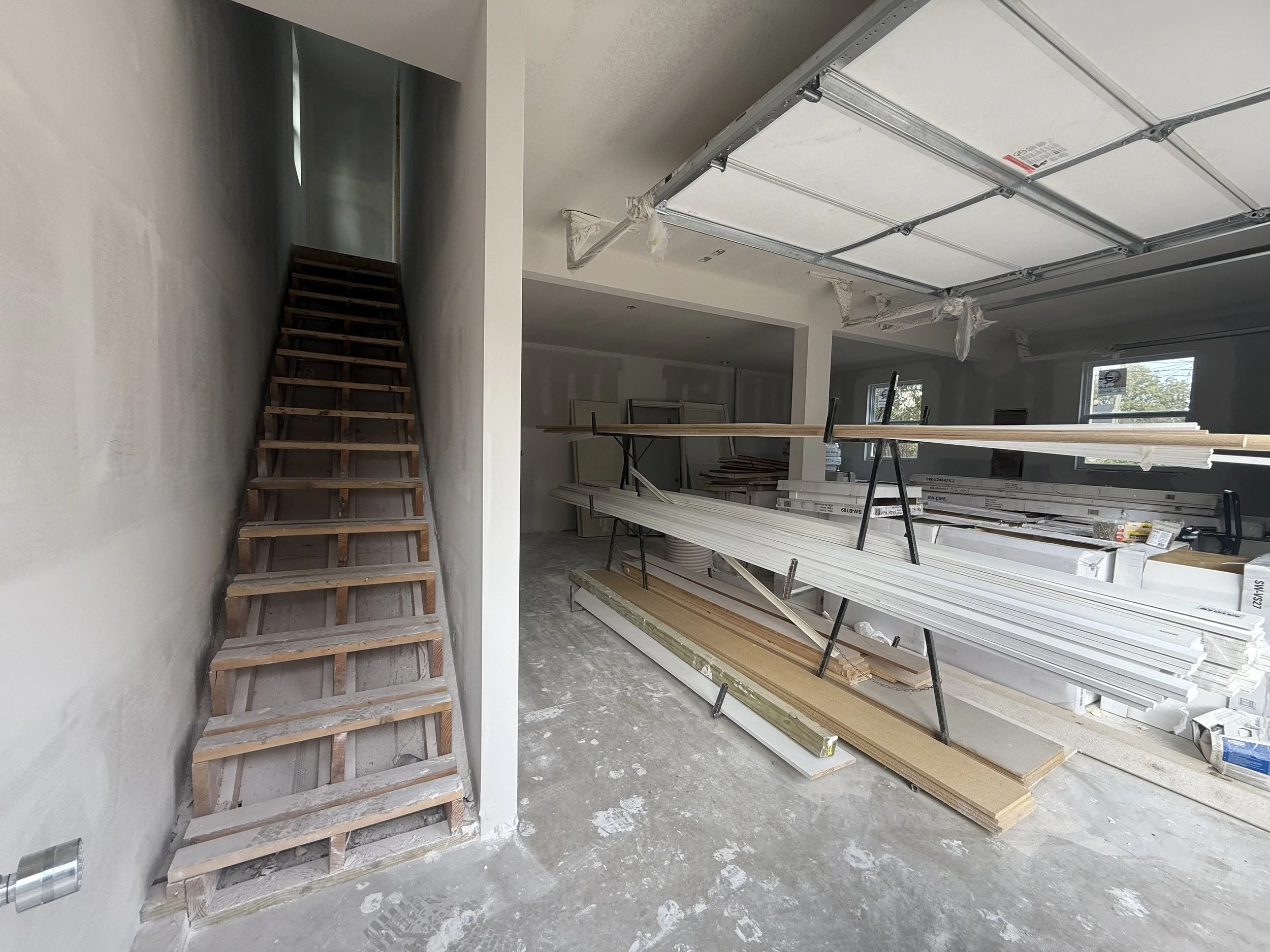 Construction site inside a house with unfinished staircase on the left, construction materials and long white boards on the right, and a partially installed garage door overhead.