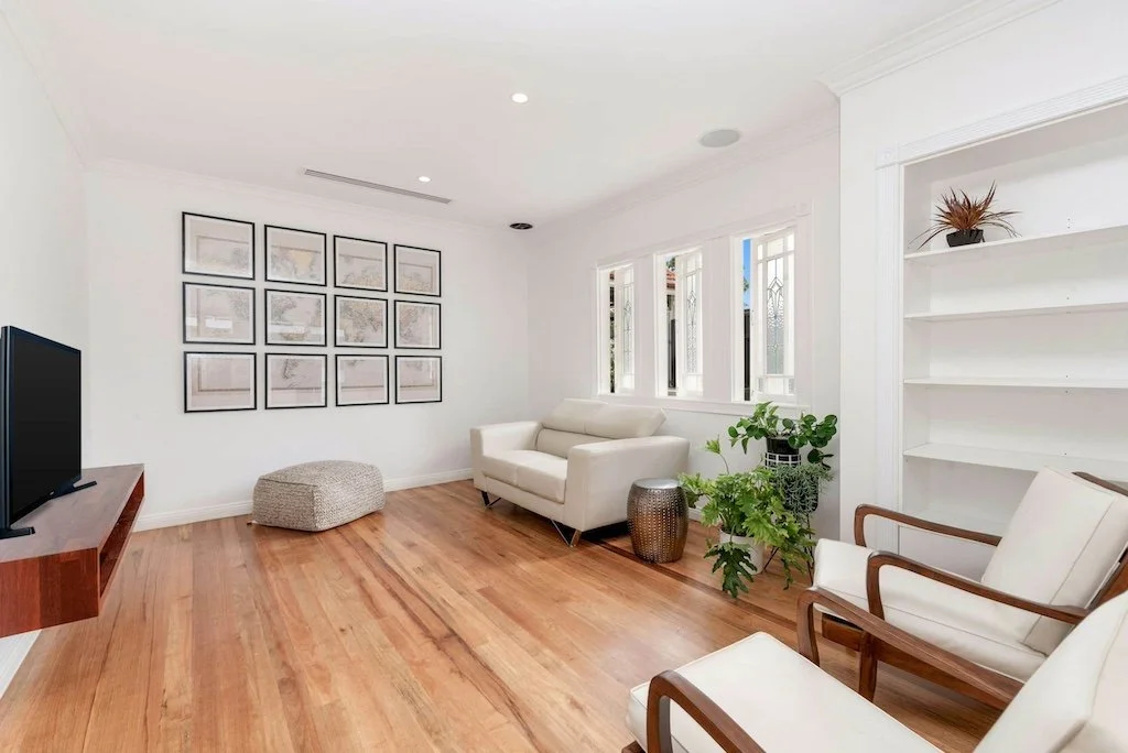 Bright living room with wood floor, white chairs, wall art, potted plants, TV on a floating shelf, and fresh interior painting.