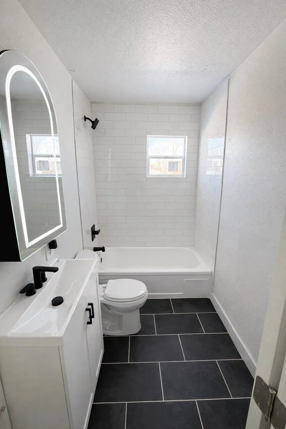Modern bathroom with black and white color scheme, featuring a white vanity with a black faucet, a wall-mounted mirror with LED lighting, a toilet, a black showerhead, and a bathtub surrounded by white subway tiles, with two small windows.
