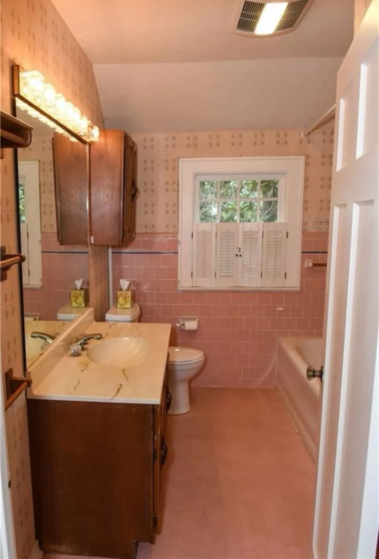 Vintage bathroom with pink tile, a single sink, a toilet, a bathtub, wooden cabinets, and a window with white shutters.