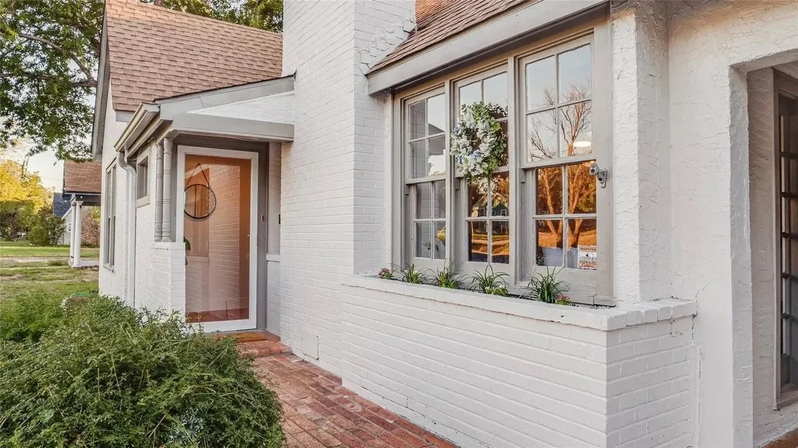 Front view of a white brick house with large windows, a small porch, and a brick walkway surrounded by greenery.