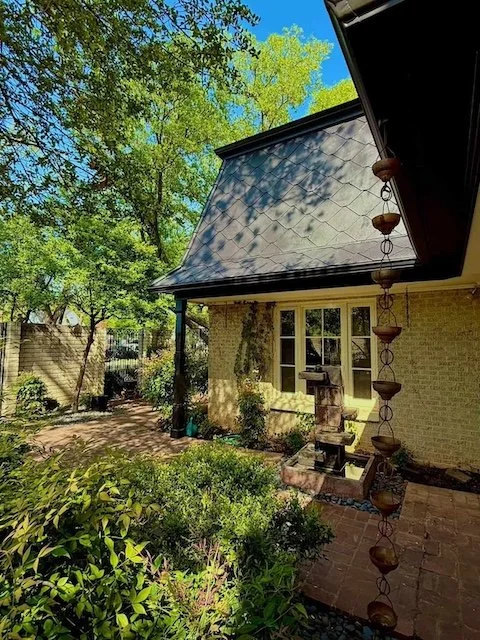 A backyard garden with lush green trees, shrubs, and a small brick patio. A decorative water fountain is present near the house wall with a window, and a hanging water feature with small bowls is hanging beside the house. The sky is bright blue with sunlight creating shadows.