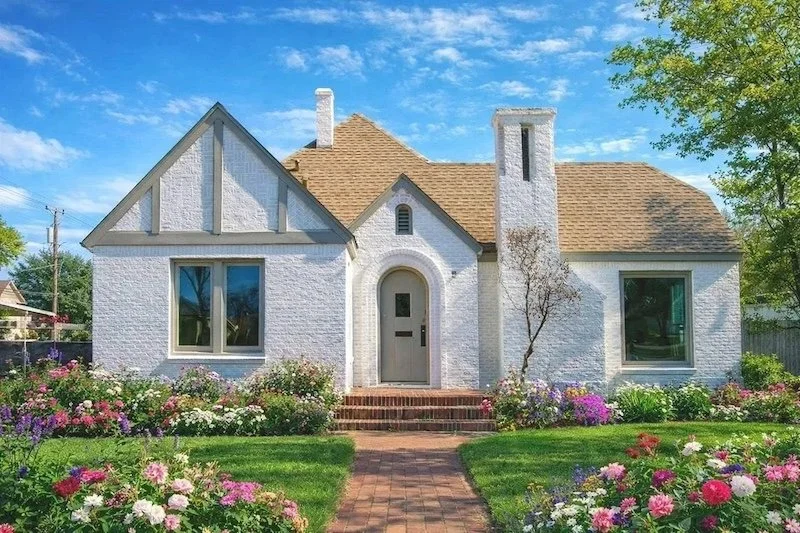 The exterior of the front of a white brick house with luscious green grass and flowers blooming