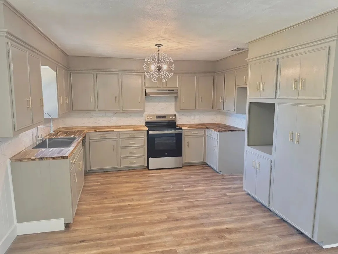 Empty kitchen with beige cabinets, wooden countertops, black stove, white marble backsplash, and wooden flooring, with a chandelier hanging from the ceiling.