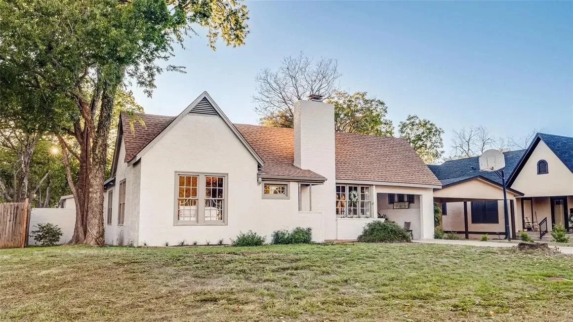 A white house with a gable roof and a chimney, with trees in the background and a lawn in the foreground. There is a neighboring house with a basketball hoop and a porch.