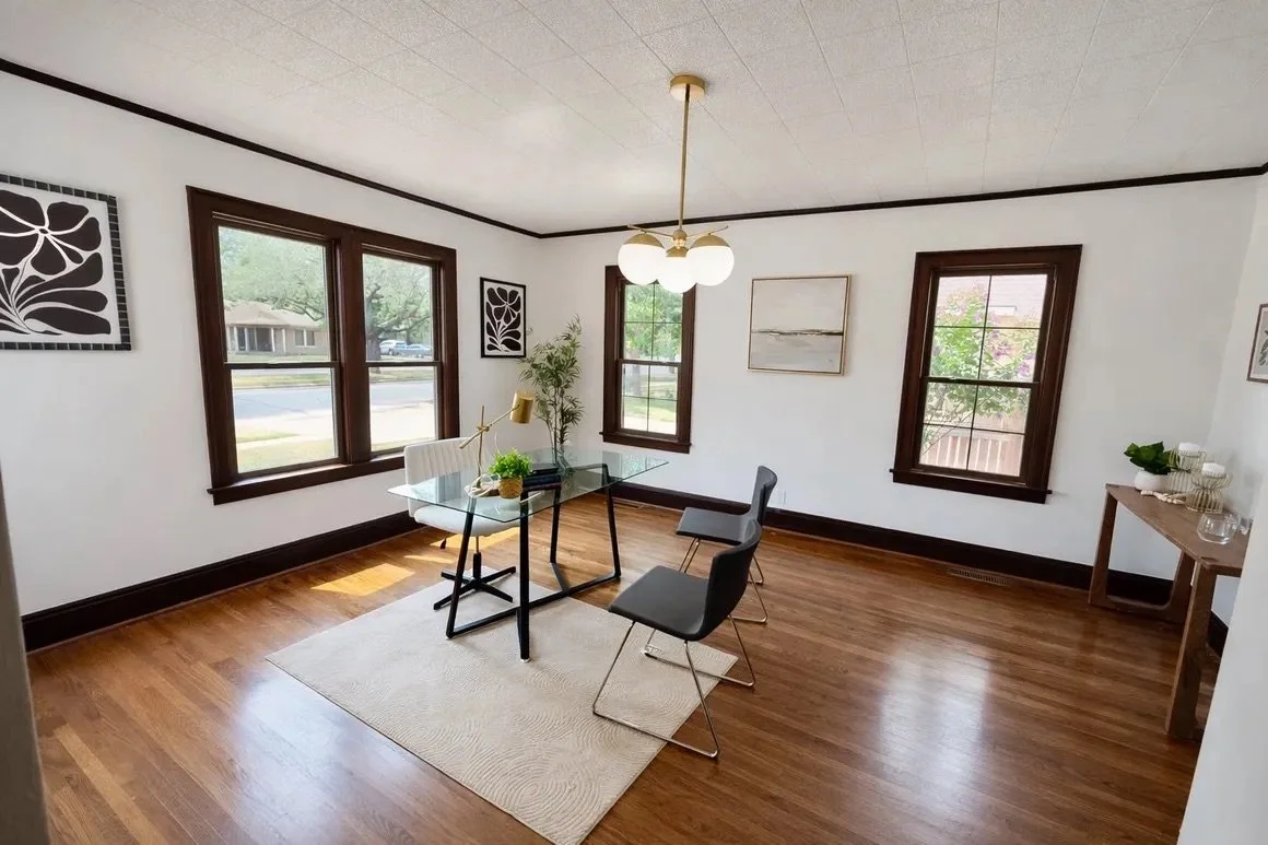 Dining room with wooden floors, white walls, three windows with dark wood trim, a modern glass dining table, four chairs, a hanging light fixture, and wall art.