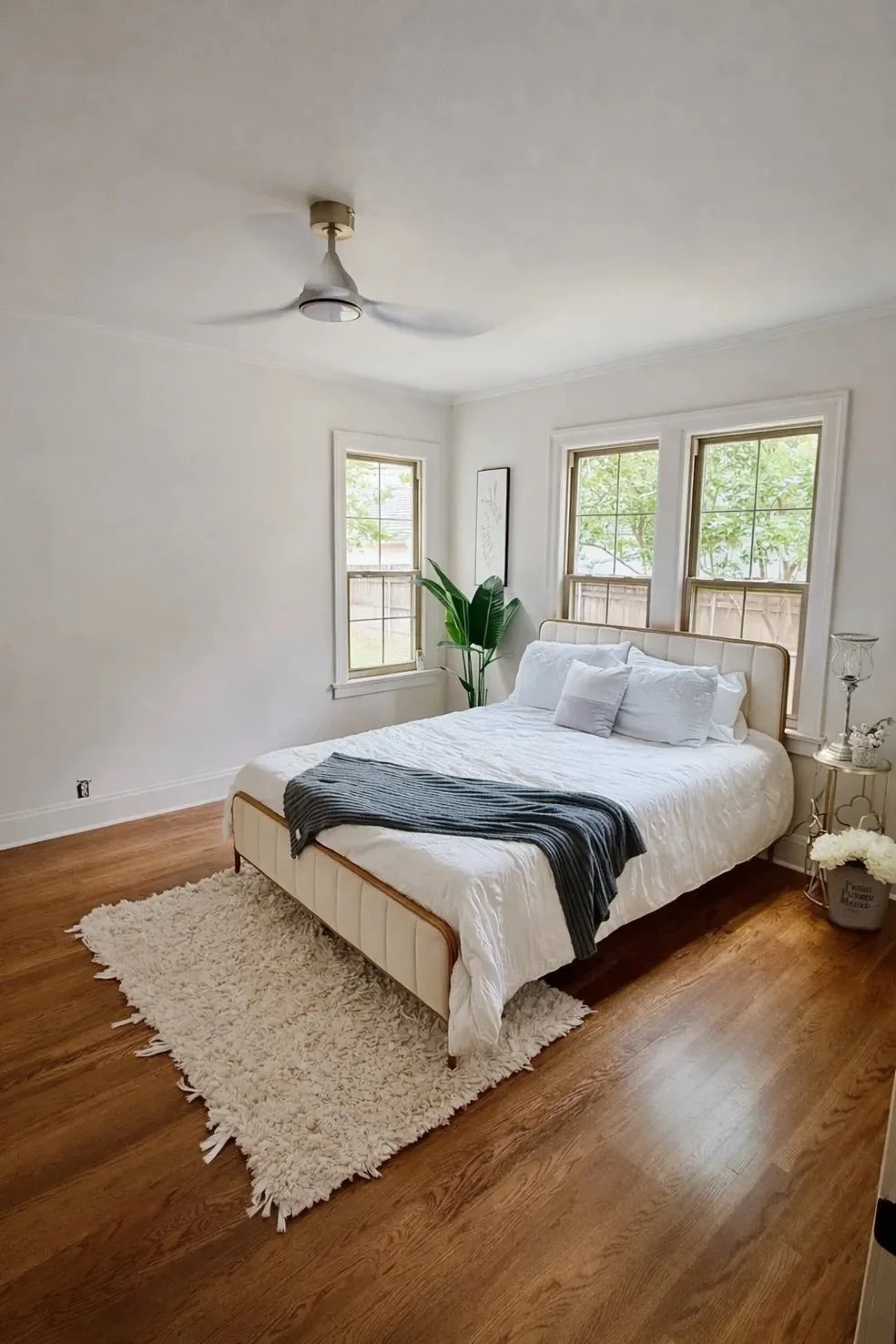 A minimalist bedroom with a white bed, multiple pillows, a gray throw blanket, a white shag rug, wooden floor, a potted plant, and two windows with green foliage outside.