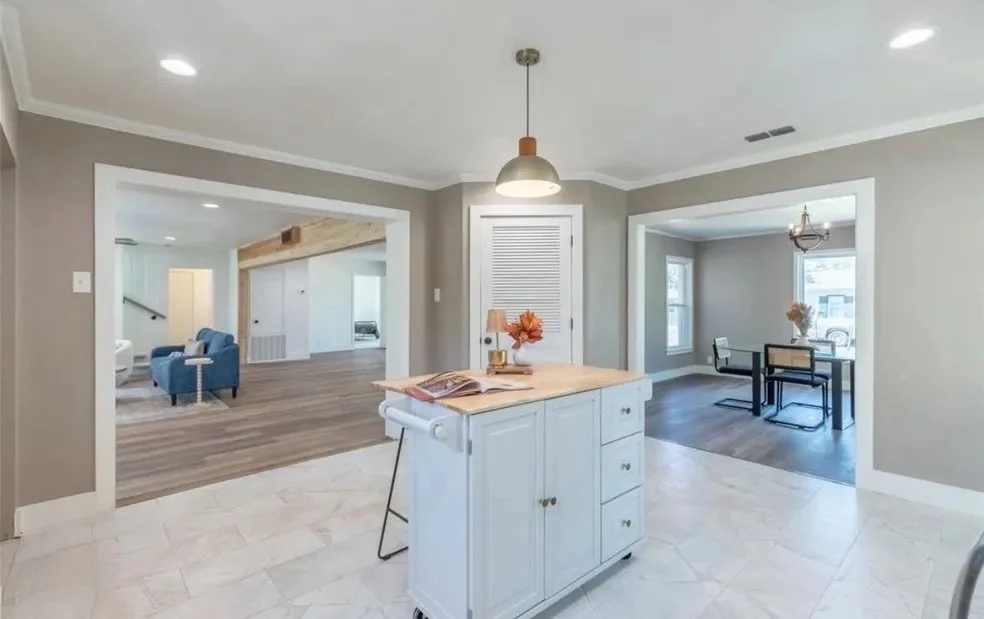Interior view of an open-plan living space with a kitchen island, hardwood floors, and a separate sitting area with a table and chairs, decorated with flowers and lamps.