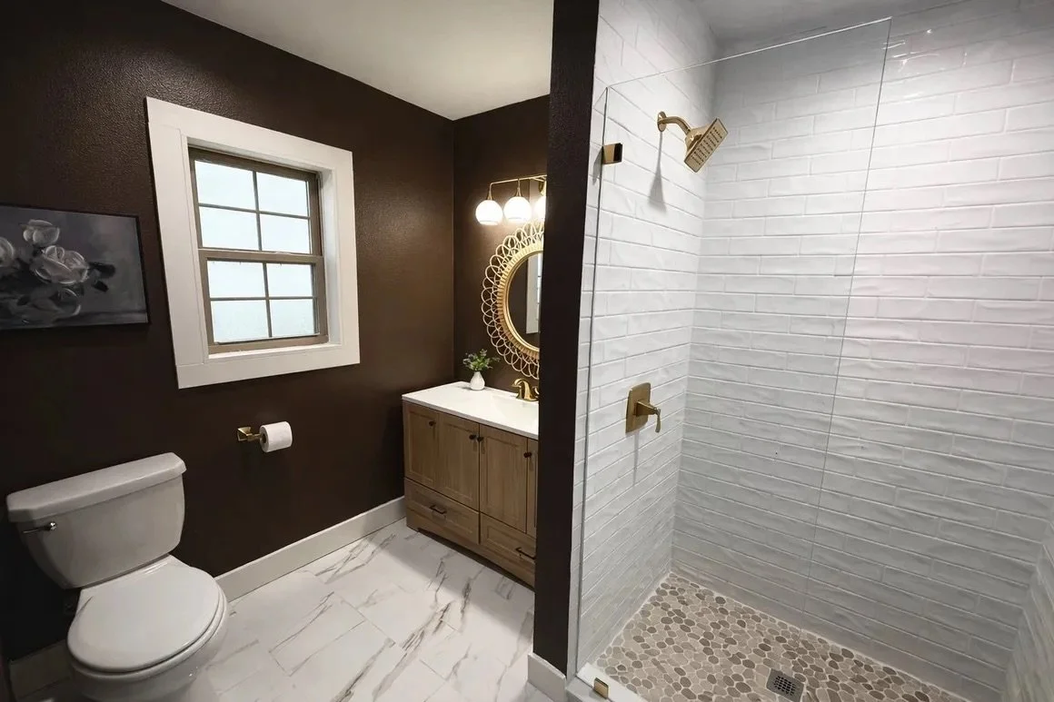 A bathroom with dark brown walls, a white toilet, a small window, a wooden vanity with a white countertop, and a decorative round mirror with lights above it. The shower area has white brick walls, a gold showerhead, and pebble stone flooring.