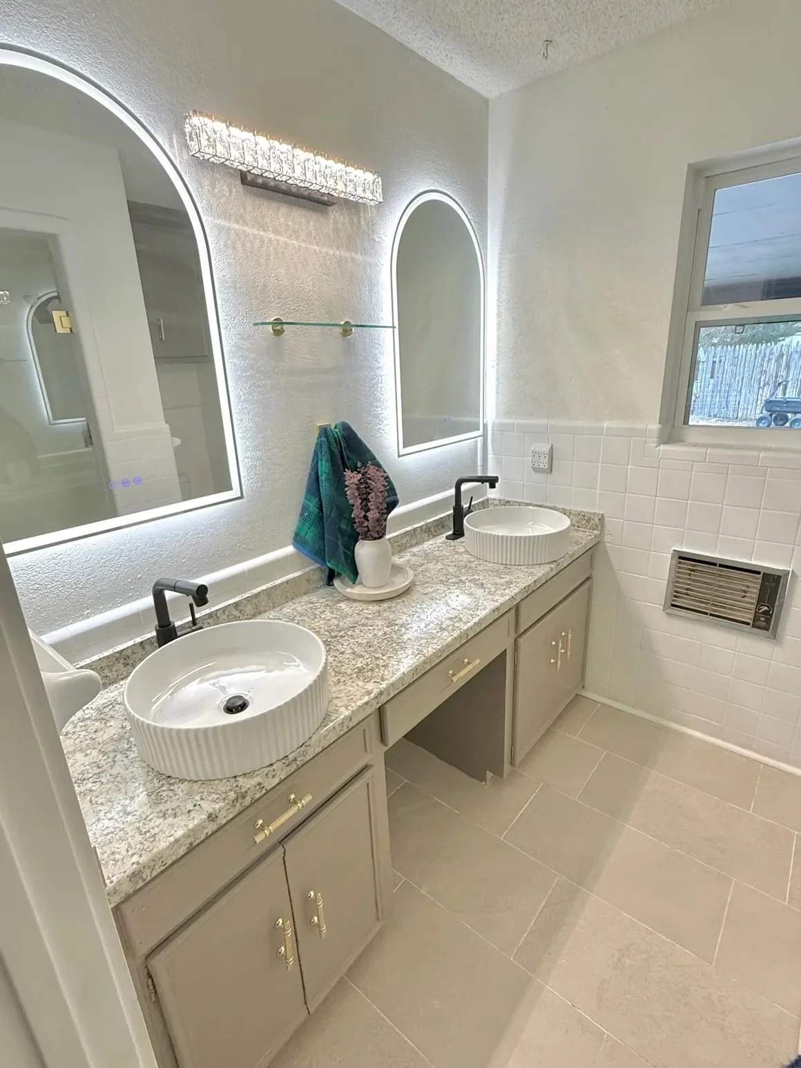 Modern bathroom with double sinks, round mirrors, and granite countertop. Blue towel and purple flowers decorate the vanity.