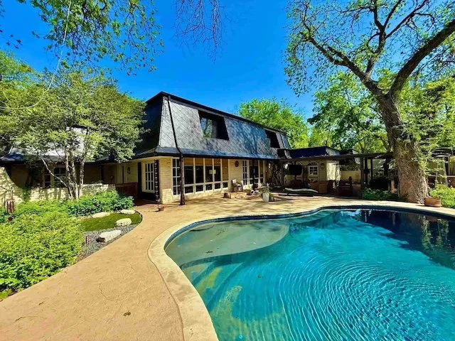Large house with dark roof and pool in backyard, surrounded by trees and lush greenery under a bright blue sky.