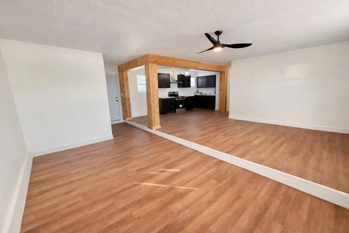 Empty living room with wood laminate flooring, white walls, a black ceiling fan, and an open kitchen area with black cabinets and a wooden support beam.