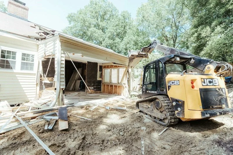A yellow JCB skid steer tackles renovation, demolishing the house side with debris scattered and exposed framing visible.