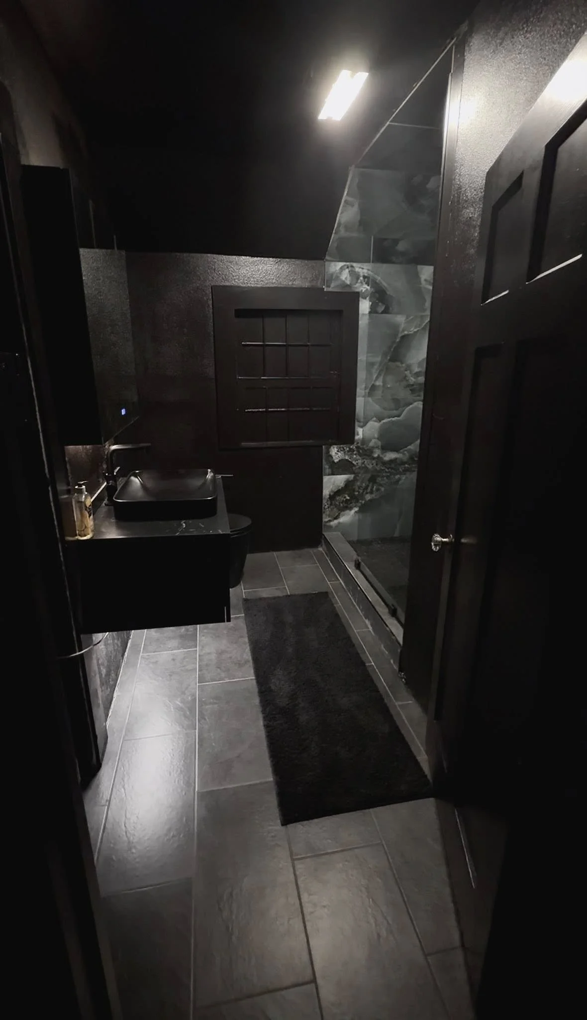 Modern bathroom with black tiles, a black sink, a dark window, a glass shower, and minimal lighting.