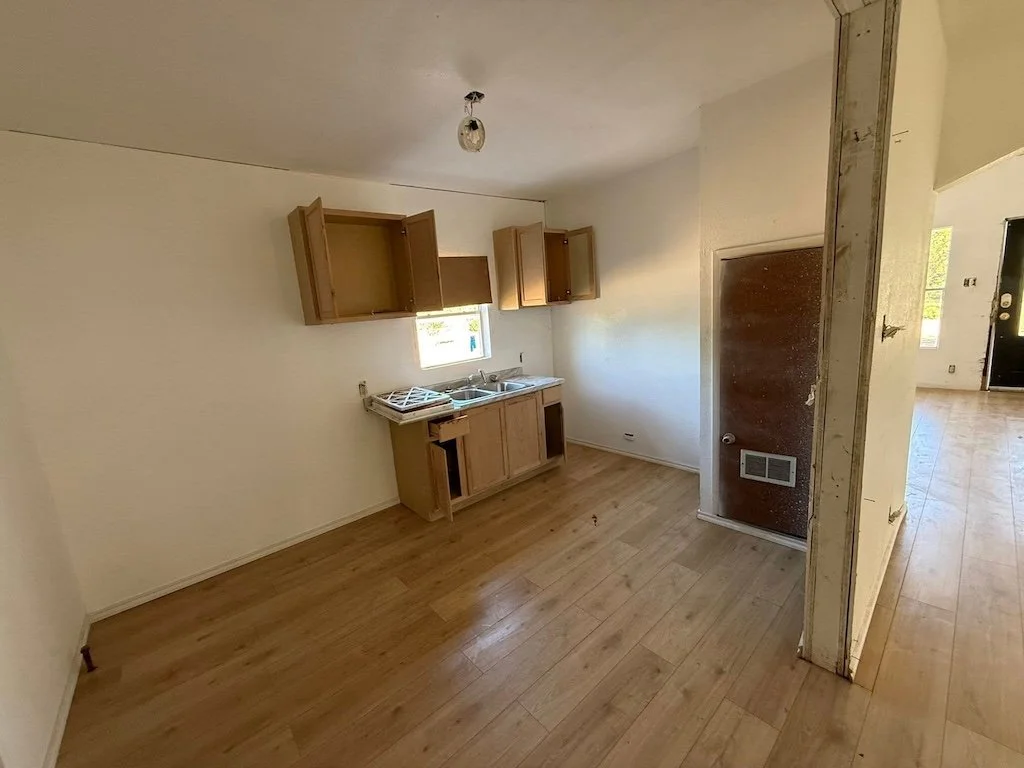 Small, empty kitchen with open cabinets, missing drawers, and a single bulb; wood floor and white walls.