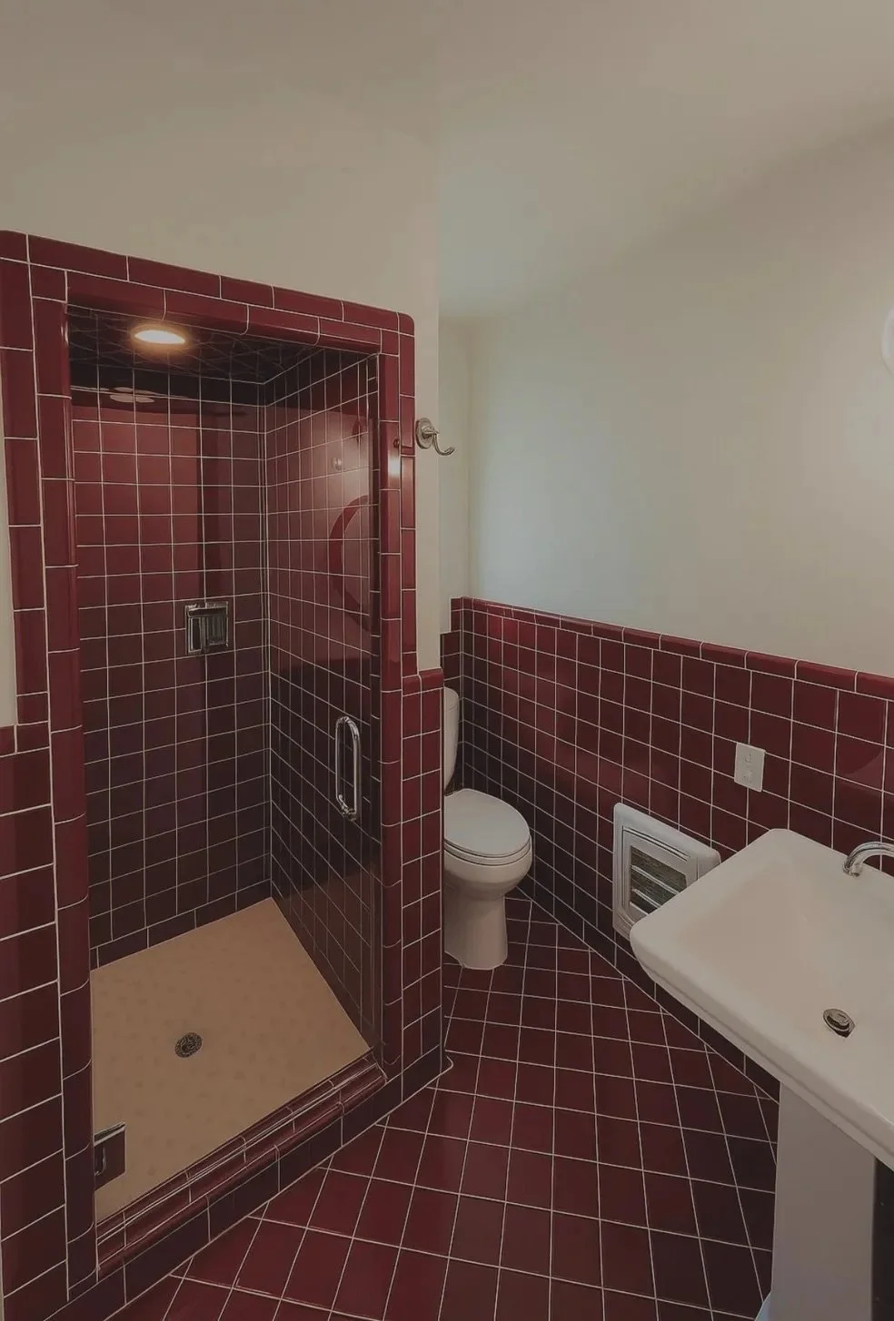 A small bathroom with a walk-in shower with red tile walls and beige flooring, a white toilet, and a white sink with a cabinet. The shower has a glass door and a ceiling light, and the room has a white vent below a power outlet on the wall.