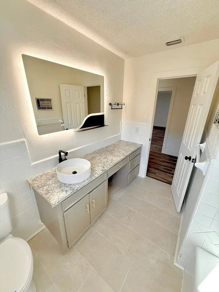 A bathroom with a granite countertop vanity, round white vessel sink with black faucet, large rectangular mirror with backlighting, a small towel rack, white tiled backsplash, beige tiled floor, and an open door leading to a room with wooden flooring.