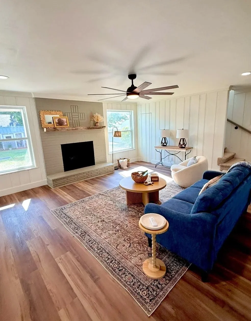 Bright living room with hardwood floors, white paneled walls, a fireplace, windows with sunlight, off-white and blue seating, a wooden coffee table, decorative lamps, and ceiling fan.
