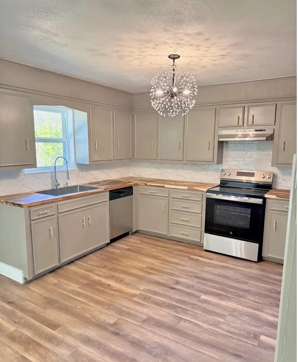 Modern kitchen with beige cabinets, wood countertops, stainless steel appliances, and a decorative chandelier in the center.