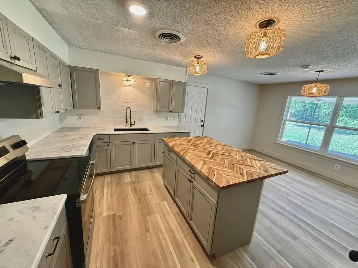 Modern kitchen with light gray cabinets, herringbone wood island, marble countertops, pendant lights, and large window.