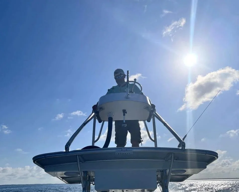 The owner of Webb Tide & Timber is standing on the upper deck of his boat, with the sun shining brightly in the clear blue sky and ocean in the background.