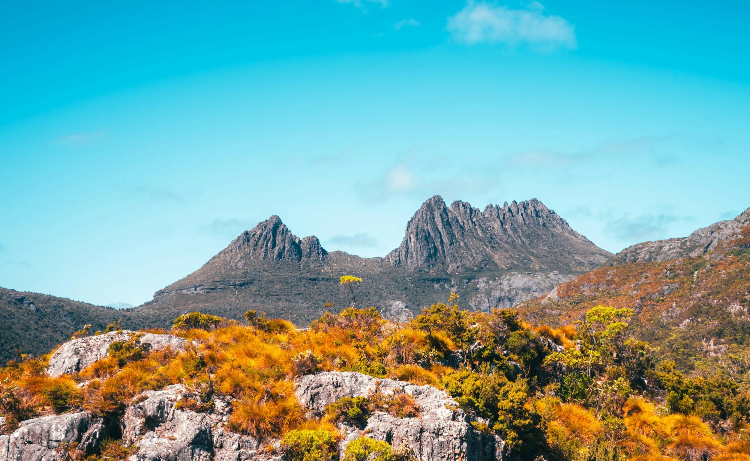 Mountain landscape with rocky peaks and autumn-colored foliage under a partly cloudy blue sky.