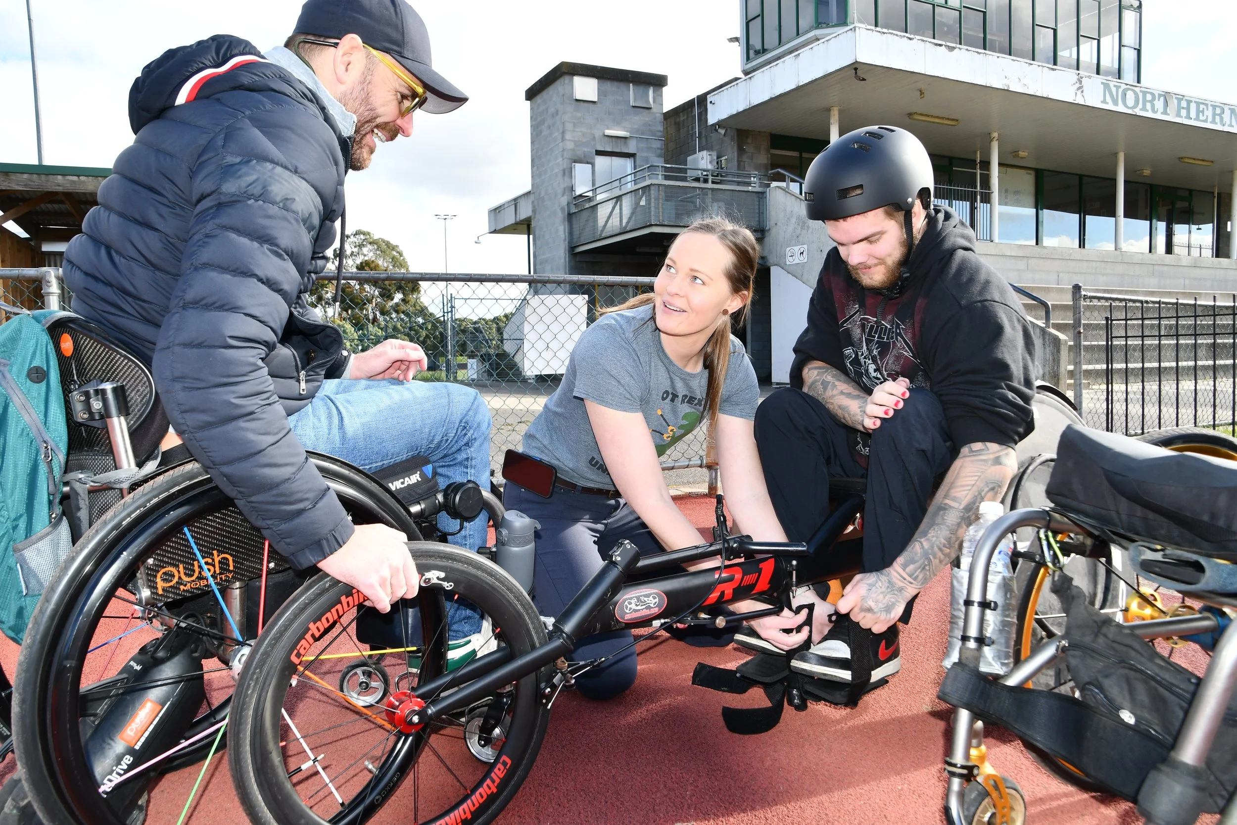 Three people, two men and one woman, are outside on a sports field, with two wheelchairs. One man is sitting in a wheelchair, while the woman is adjusting something on the other. The man in the wheelchair is wearing a black helmet and a black hoodie, and has tattoos on his arms. The woman has long brown hair and is wearing a gray T-shirt. The other man is standing, smiling, and wearing a baseball cap, glasses, a puffy jacket, and jeans. They are engaged in preparing or adjusting the wheelchairs.