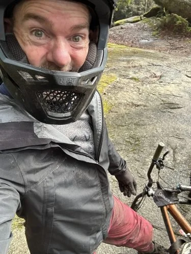 A man wearing a helmet, jacket, and riding gear taking a selfie outdoors, standing next to his orange mountain bike on a rocky trail with greenery and moss in the background.