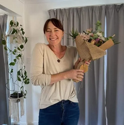 A woman with short brown hair, wearing a beige long-sleeve shirt and dark jeans, is smiling and holding a bouquet of flowers wrapped in brown paper. She is standing in a room with gray curtains and a decorative plant on a shelf in the background.