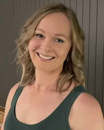A smiling woman with wavy blonde hair in a sleeveless green top standing in front of a dark wooden wall.