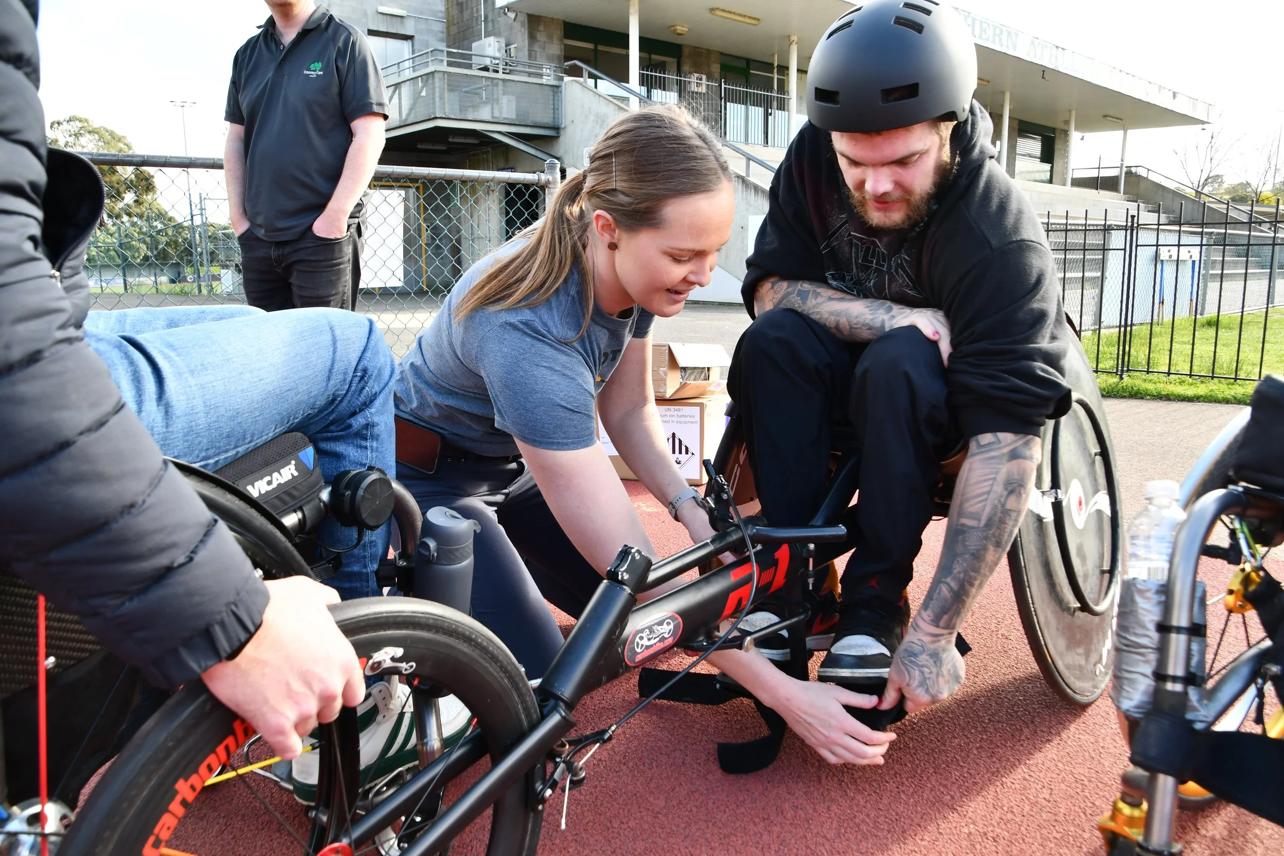 A woman in a grey t-shirt helping a man in a wheelchair attach to a black racing wheelchair. The man has a helmet and tattoos, and they are on an outdoor track. A person in a black jacket is standing nearby, and there is another wheelchair on the right side of the image.