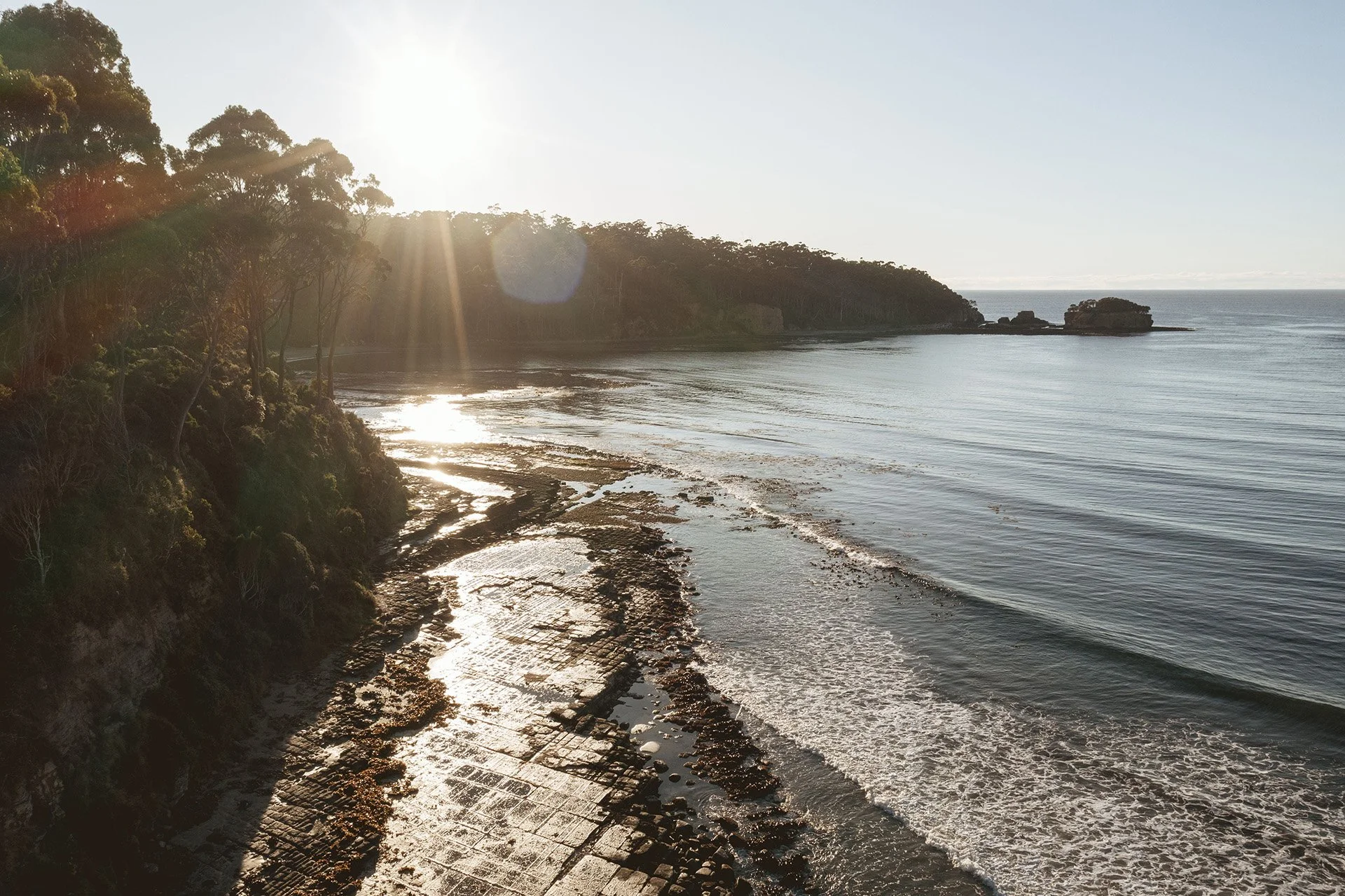 Sunset over a rocky coastline with trees on the left, gentle waves on the shore, and a small rock formation in the water on the right.