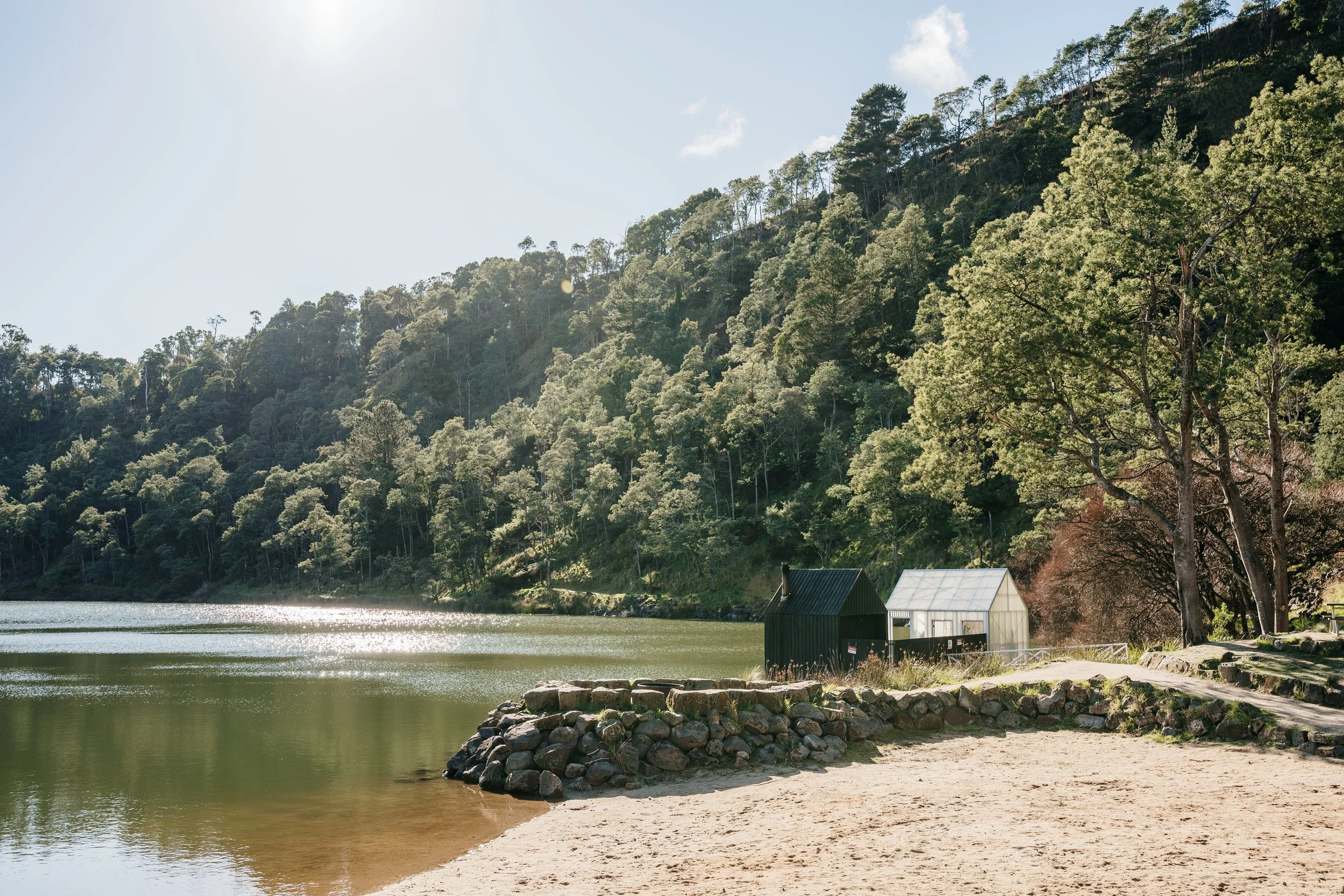 A lakeside scene with sandy beach, small stone pier, two small cabins, one dark and one white, and a forested hillside in the background under a clear sky.
