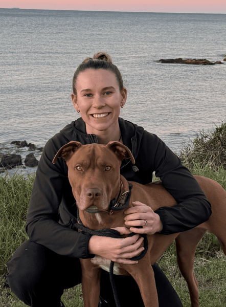 A woman smiling and holding a brown dog near a body of water at sunset.
