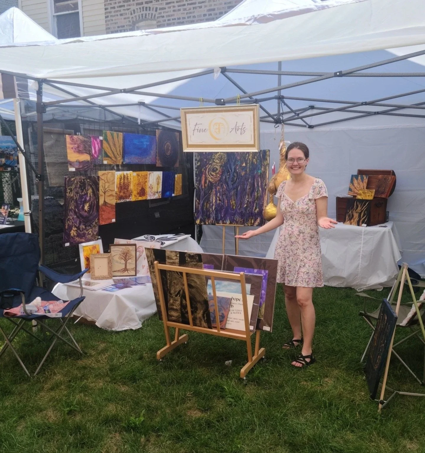 A veteran artist standing inside an art booth at an outdoor event, smiling with her hands gesturing towards her artwork. The booth displays her paintings, a sign reading ‘Fine Arts,’ and art pieces on tables and walls.