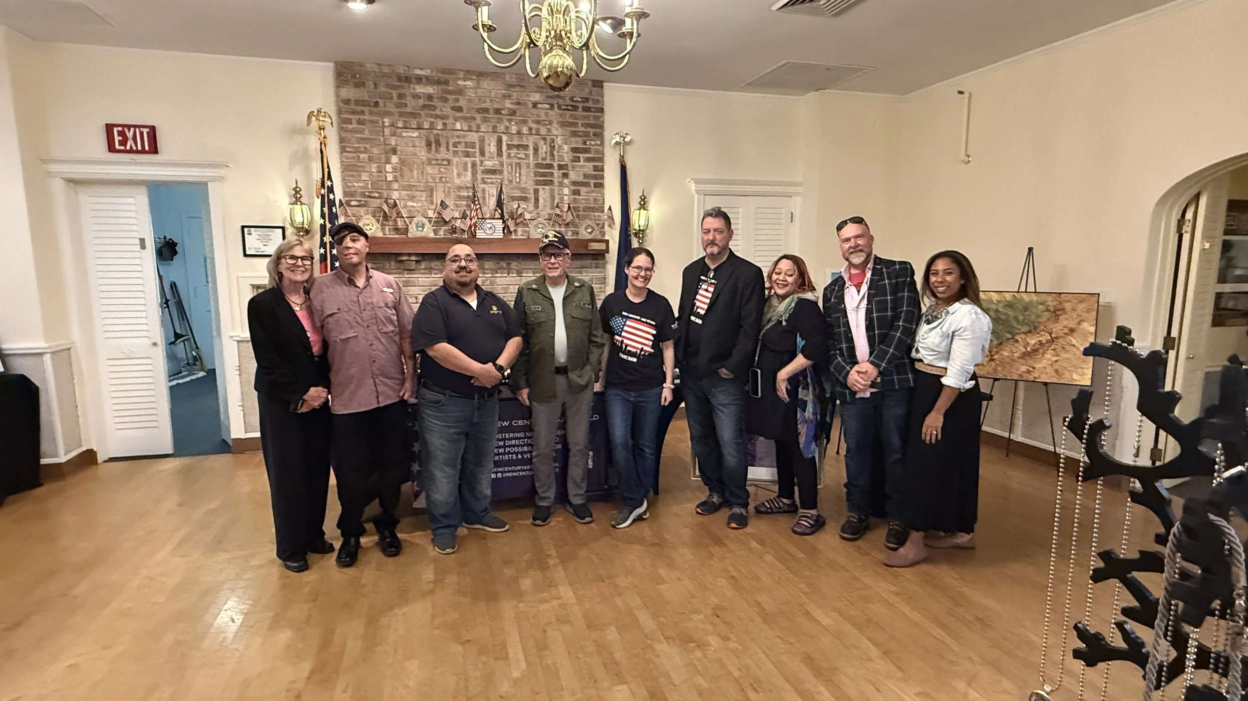 Group of nine veterans standing together inside a room with a brick fireplace, American flags, and a chandelier, posing for a photo.