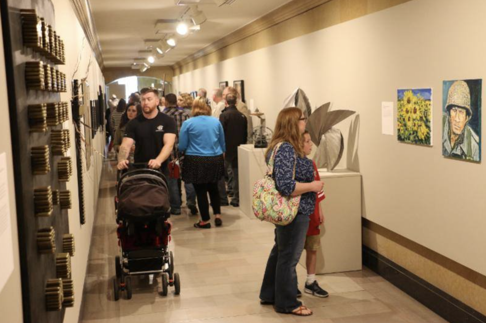 People viewing artwork in an art gallery, including a woman with a colorful bag and a young boy, with various paintings and sculptures on the walls and sculptures on display.