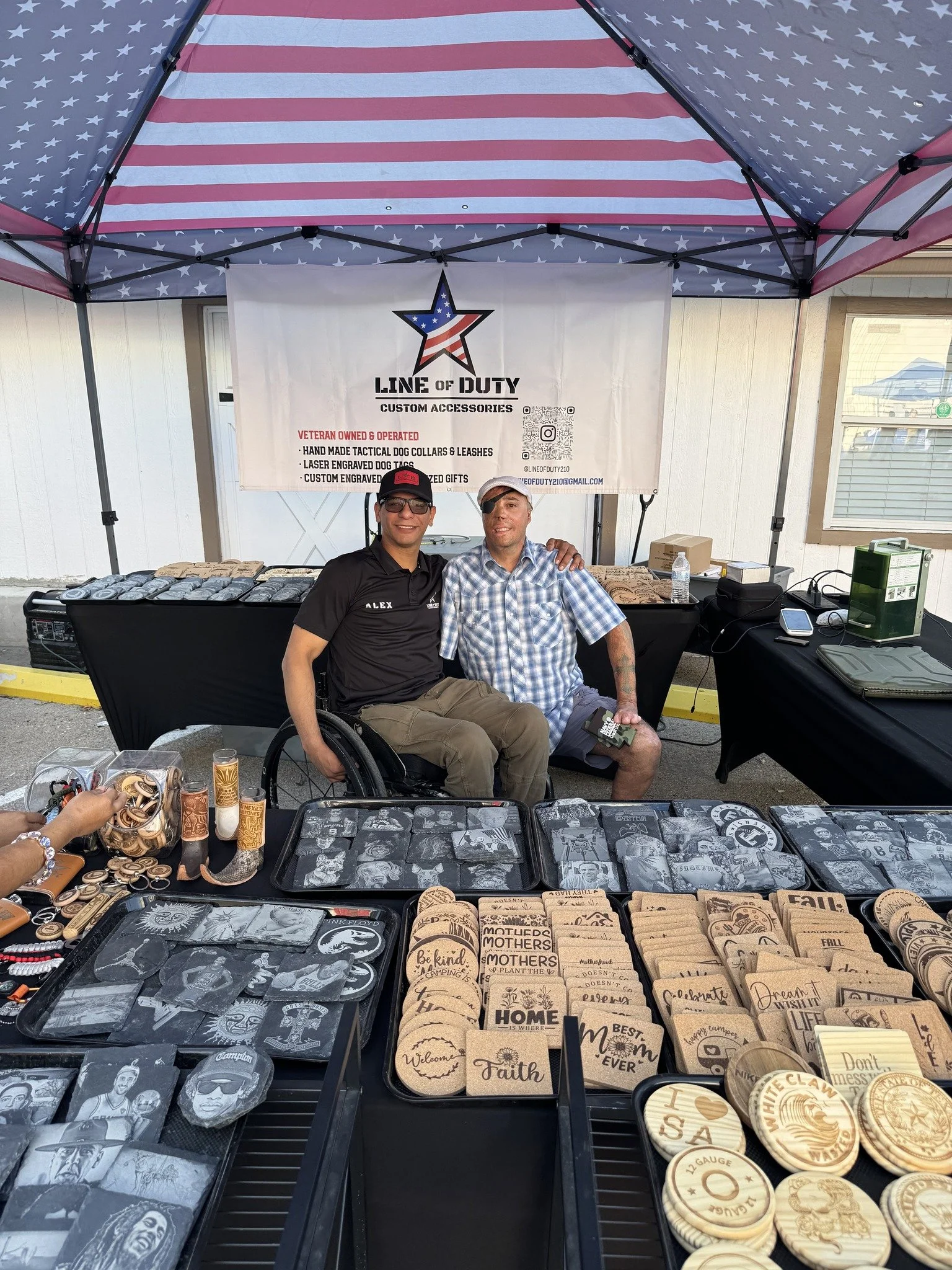Two veterans sitting together at an outdoor market stall under a patriotic umbrella. The stall displays engraved accessories, including coasters and patches, with a banner advertising veteran-owned custom accessories.