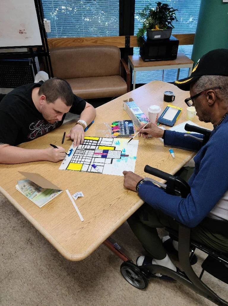 Two  veterans are sitting at a wooden table playing a bingo game with markers. One person is in a wheelchair. They are indoors, near a window with blinds, with a background that includes a couch, a potted plant, and a microwave.