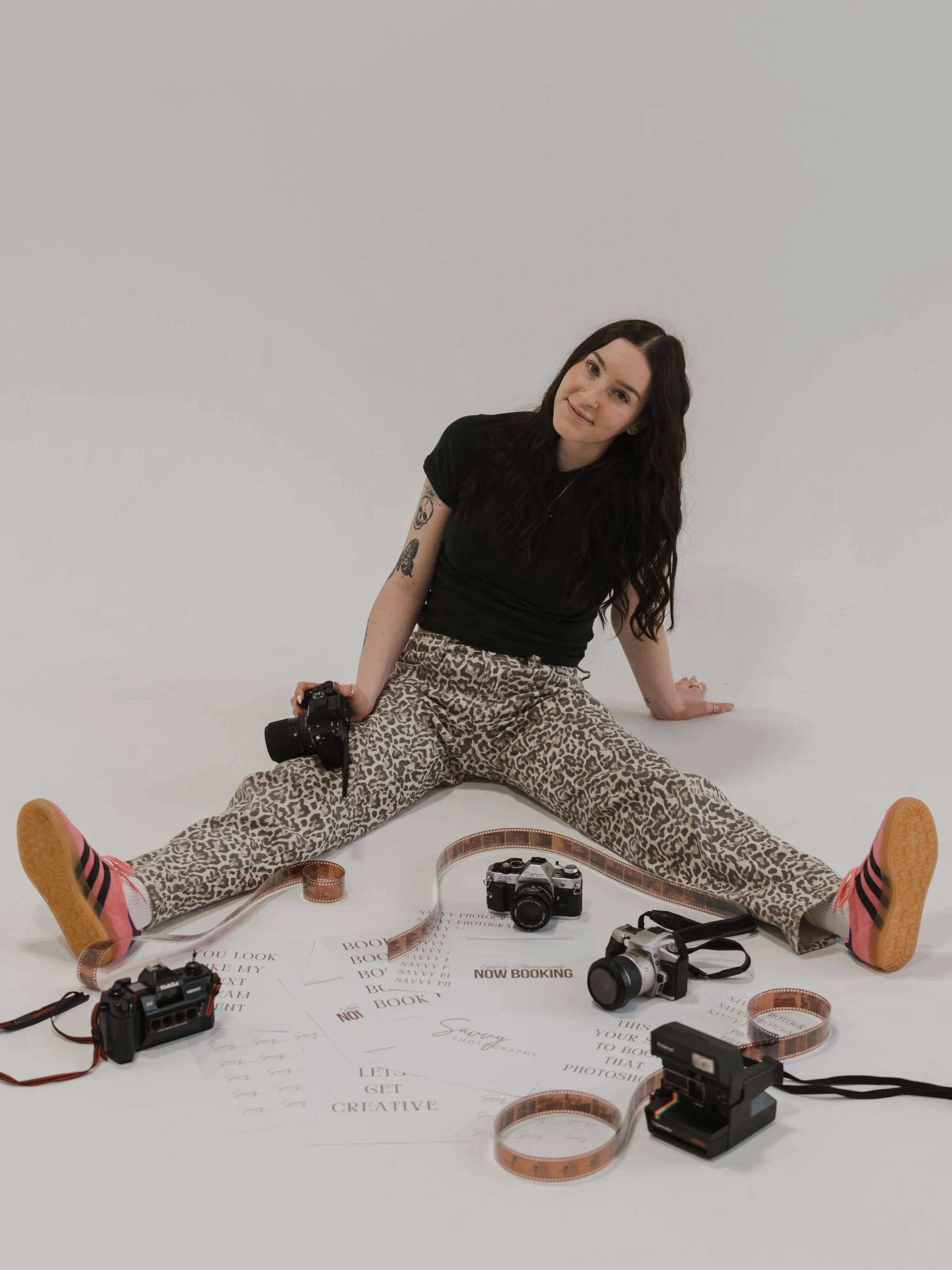 A woman sitting on the floor with her legs spread apart, wearing leopard print pants and a black shirt, surrounded by photography equipment and film strips, with a white background. Branding photos at the Opal Room Collective in Rapid City, SD.