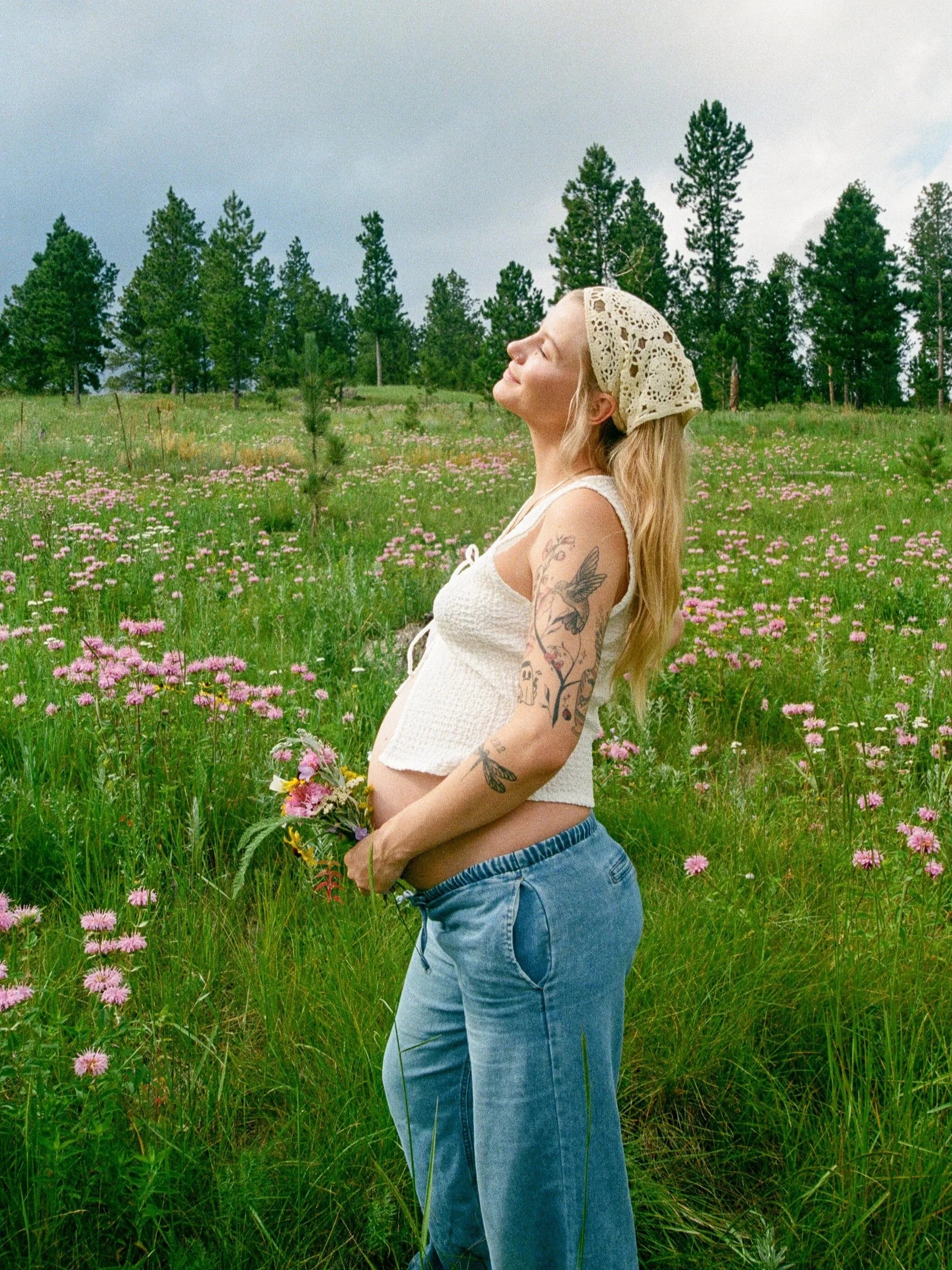 A woman with tattoos on her arm and a flower crown stands in a meadow of pink flowers with her eyes closed and a serene expression, holding a small bouquet.