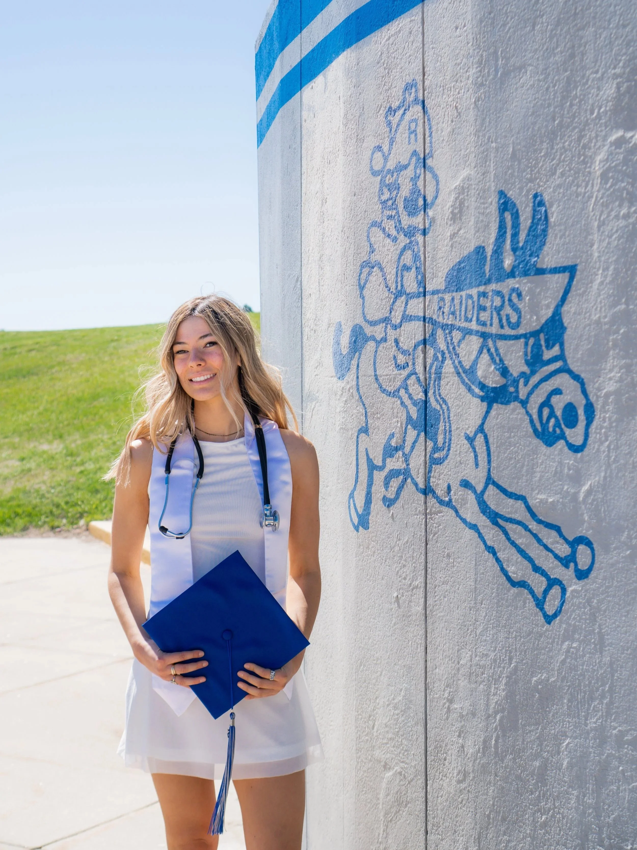 A young woman in a white dress holding a blue graduation cap, standing outdoors next to a wall with a painted logo of a knight on a horse with the word 'Raiders' on a shield (Rapid City Stevens High School), under a clear blue sky and green grass.