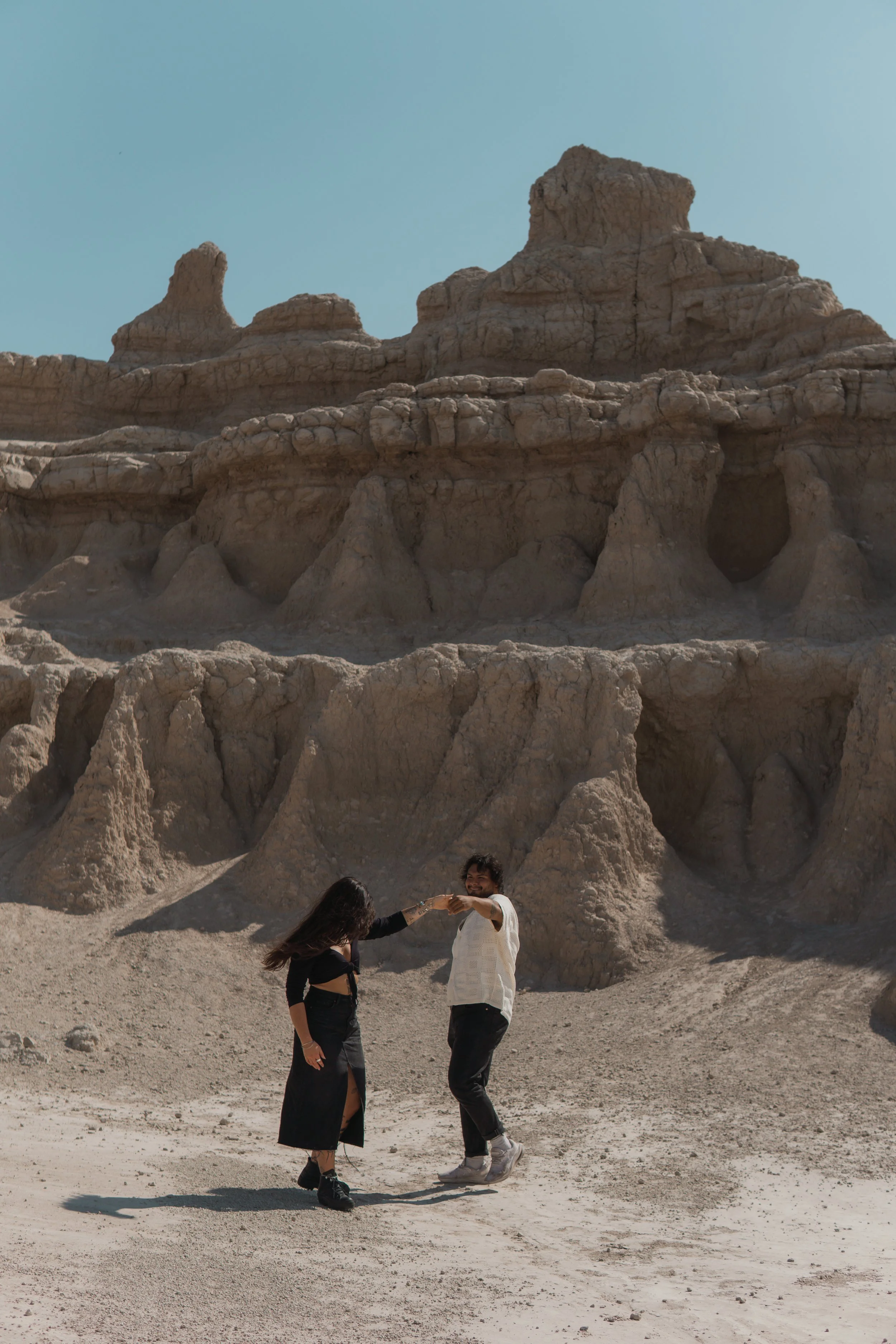 A young man and woman dance together in the Badlands landscape with large rock formations in the background. The woman wears a black dress with a slit, and the man wears a white shirt and black pants. The sky is clear and blue.