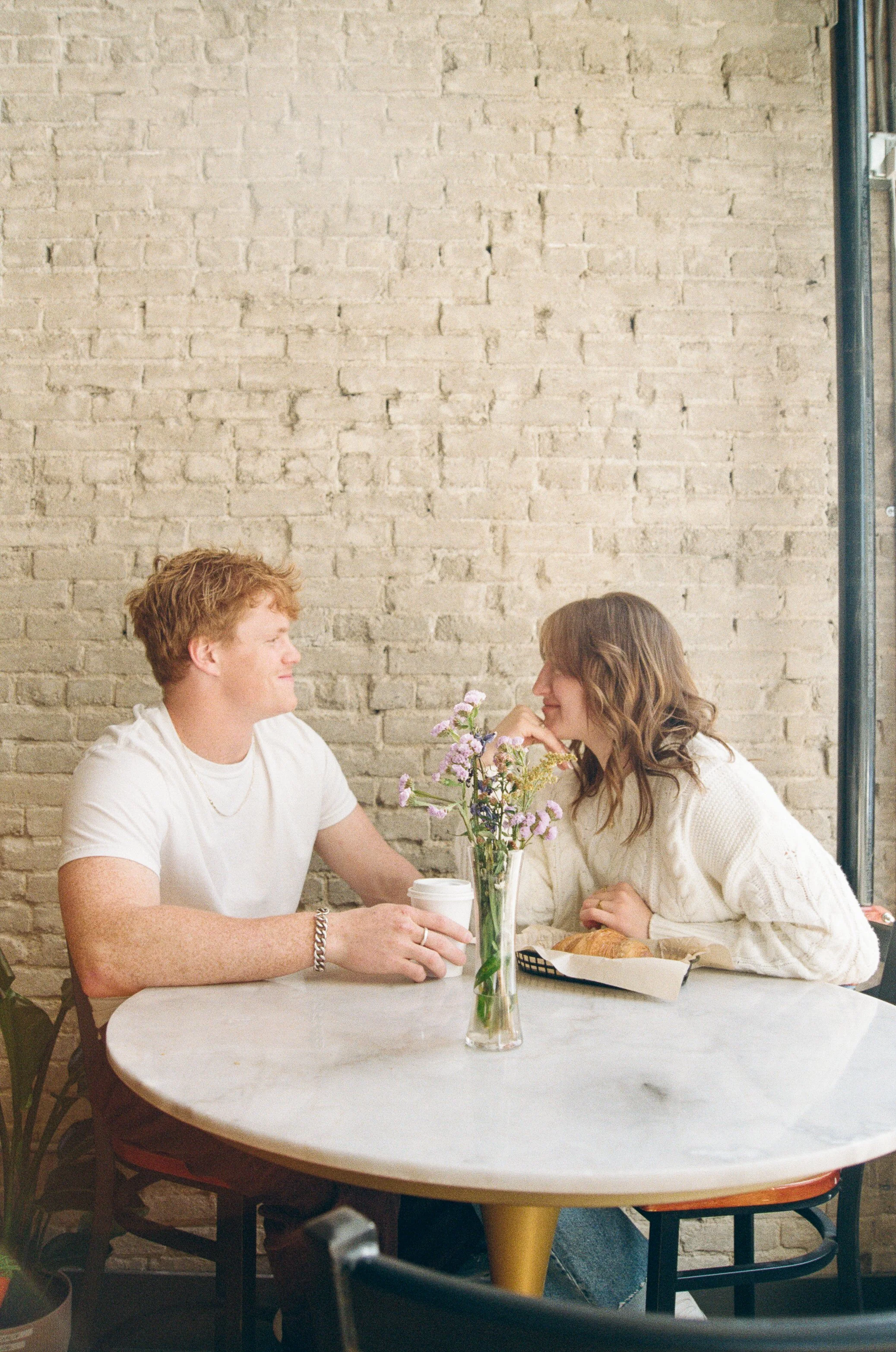 A young man and woman sitting at a round marble table in a cafe, facing each other with a flower arrangement in between. The man is smiling, holding a white takeaway coffee cup; the woman is leaning forward, smiling, with a croissant on a tray in front of her. They sit against a brick wall, with a plant in the foreground.