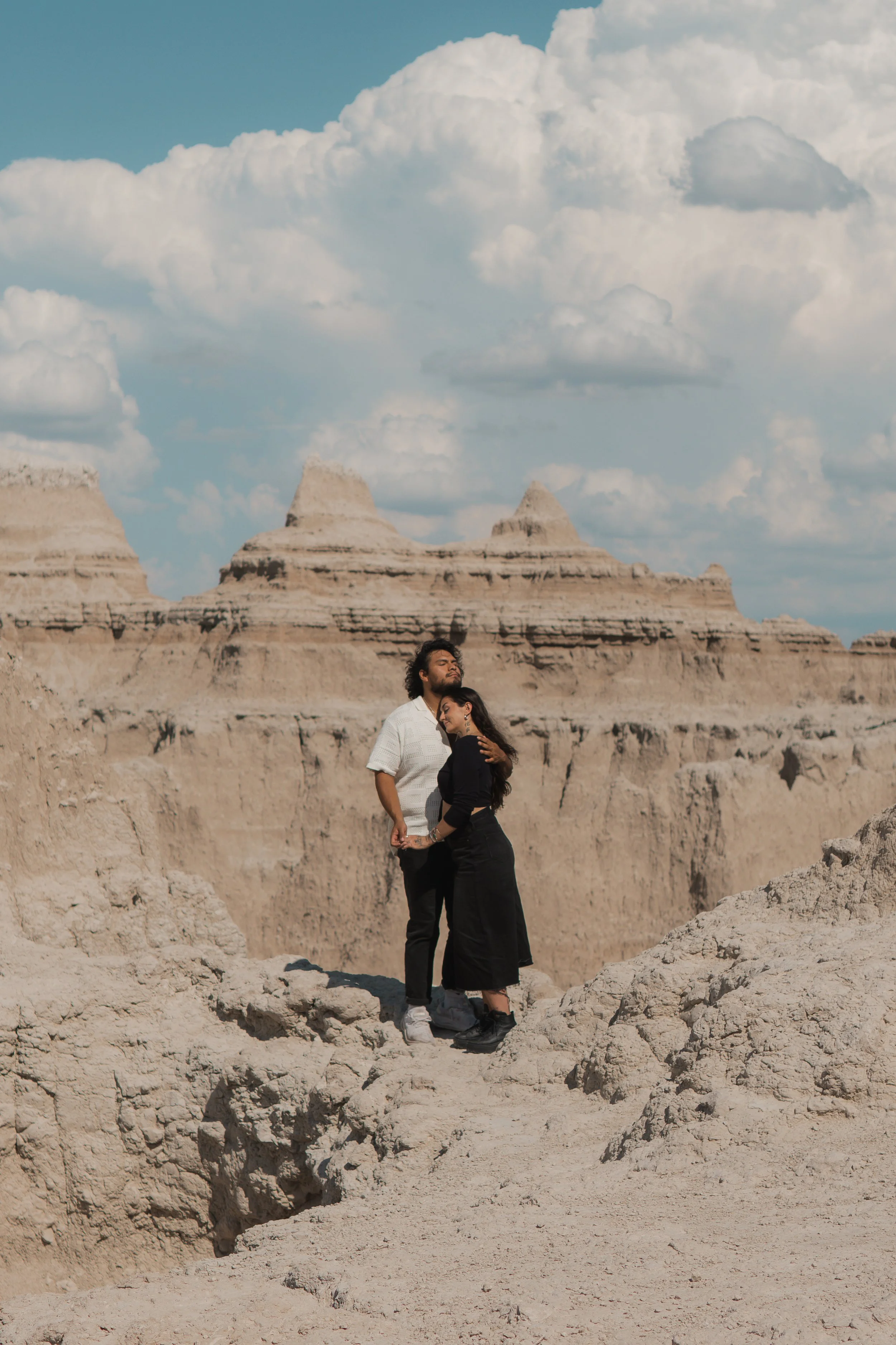 A couple standing close together in a desert landscape with eroded rock formations and a cloudy sky in the background.