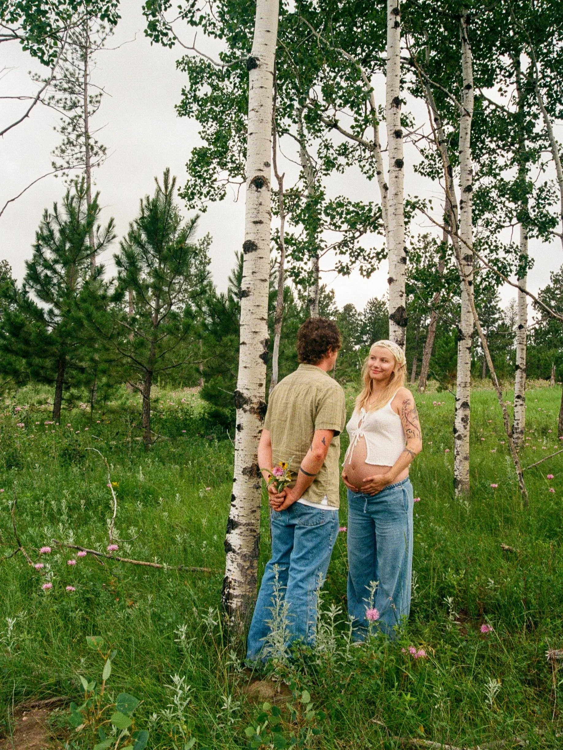 A pregnant woman and a man standing in a forest with green trees and grass, the woman is smiling and cradling her pregnant belly, while the man is holding a small bouquet of flowers behind his back. Cutest couples maternity photos.